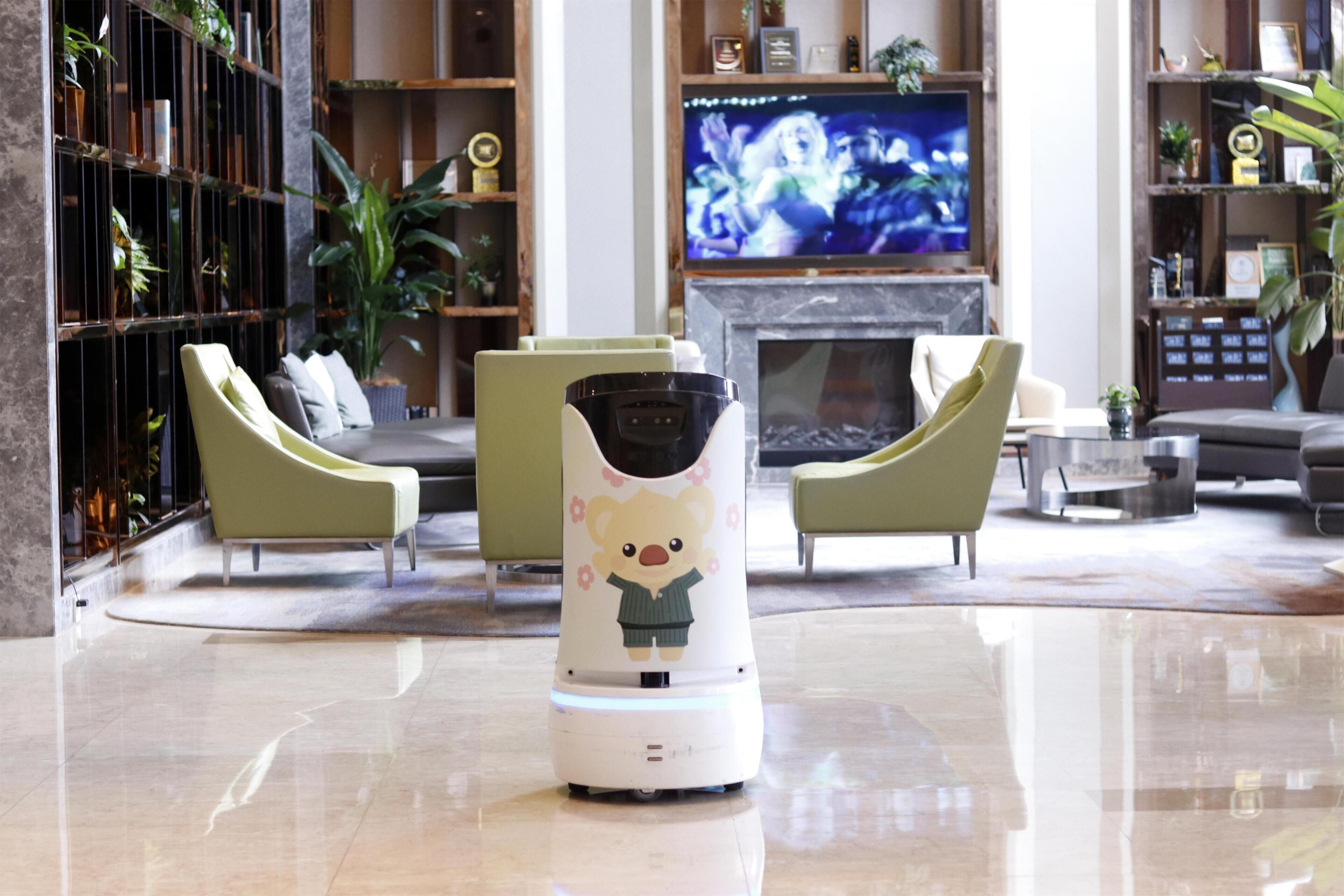A robot in a modern lobby with green sofas, a fireplace, and a TV.