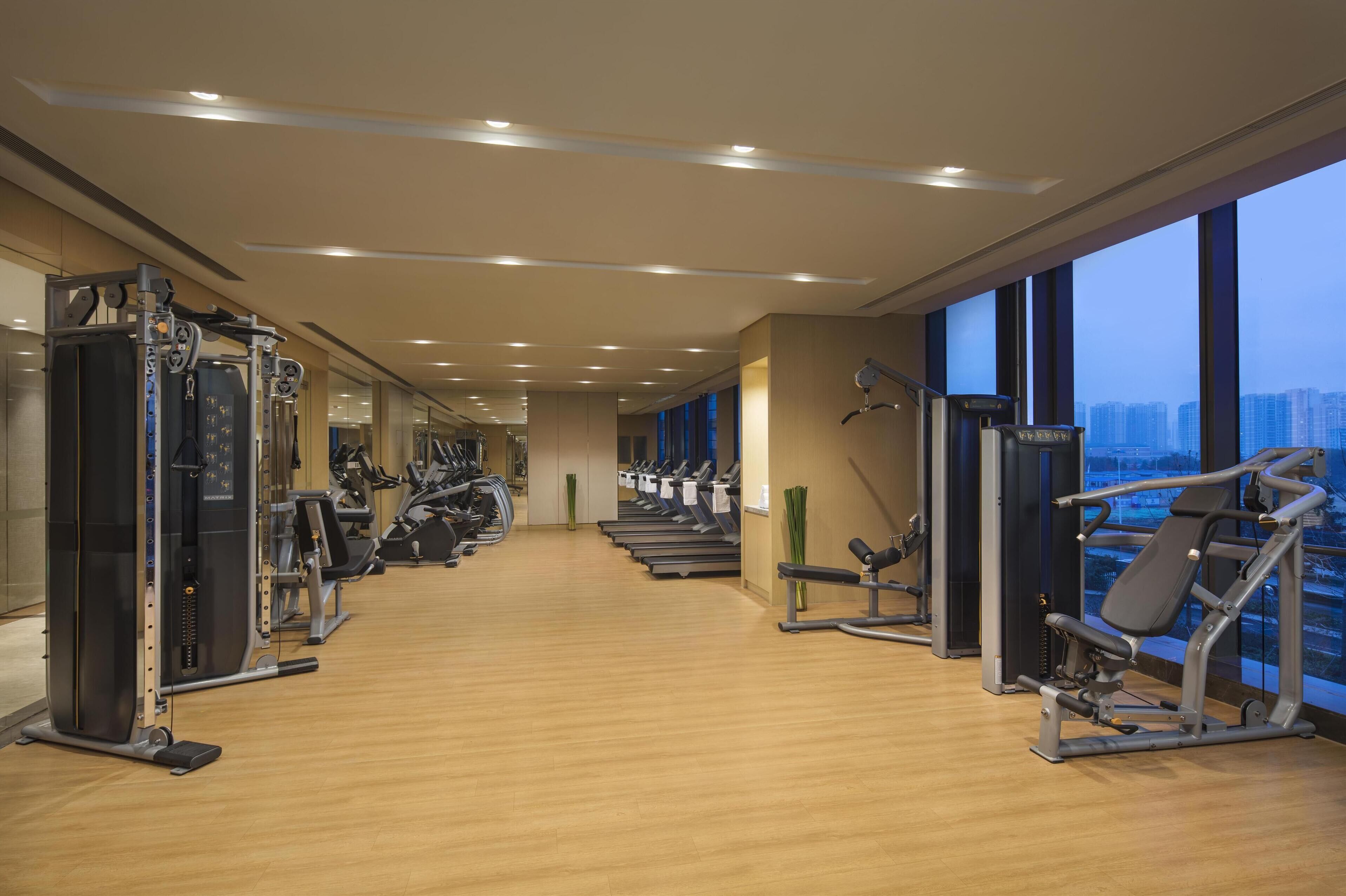 Uma academia moderna com equipamentos de cardio, máquinas de peso e uma grande janela com vista para a cidade.