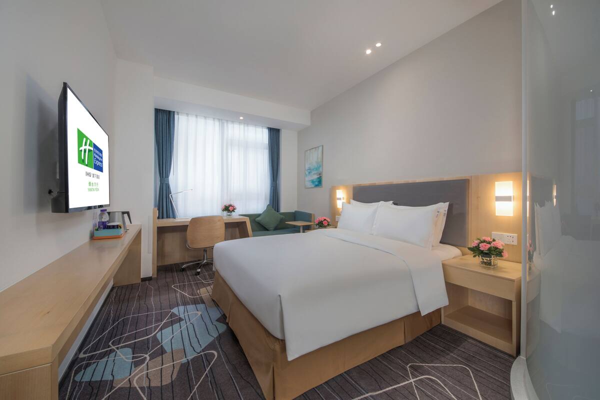 A hotel room with a large bed, a desk, a chair, a TV, and a window with curtains.
