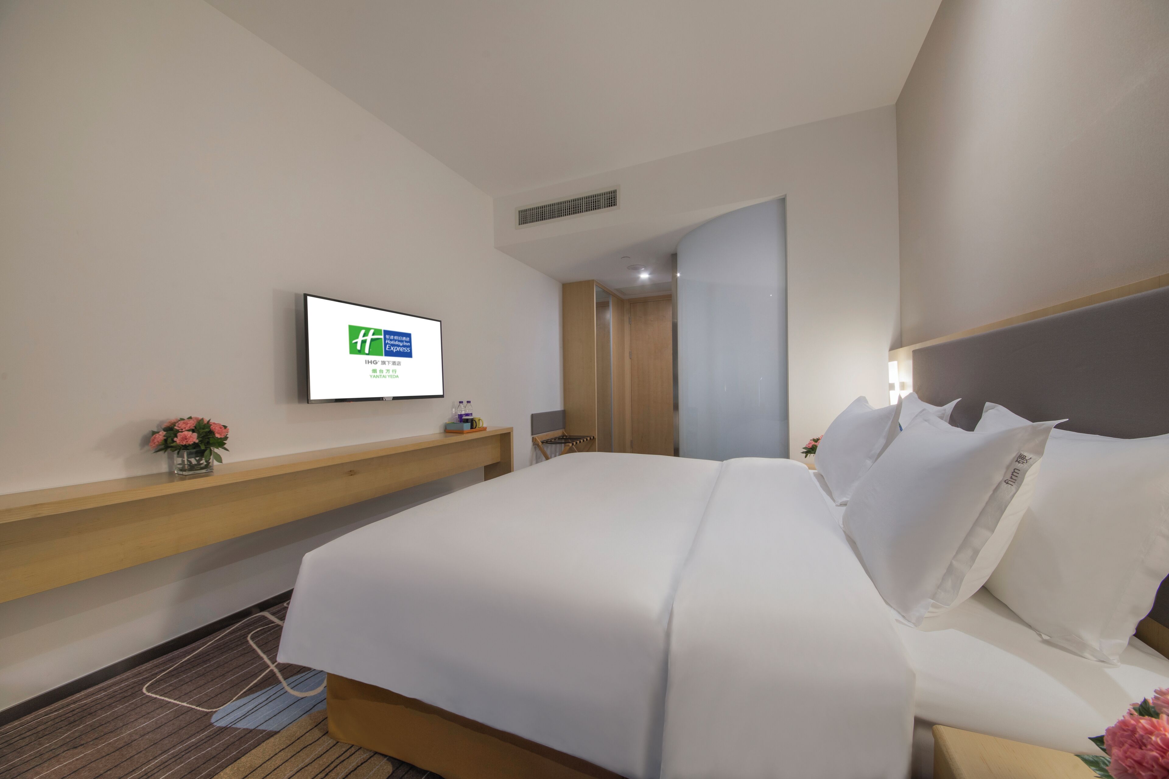 A hotel room with a bed, a TV mounted on the wall, a wooden nightstand, and a small vase with flowers.