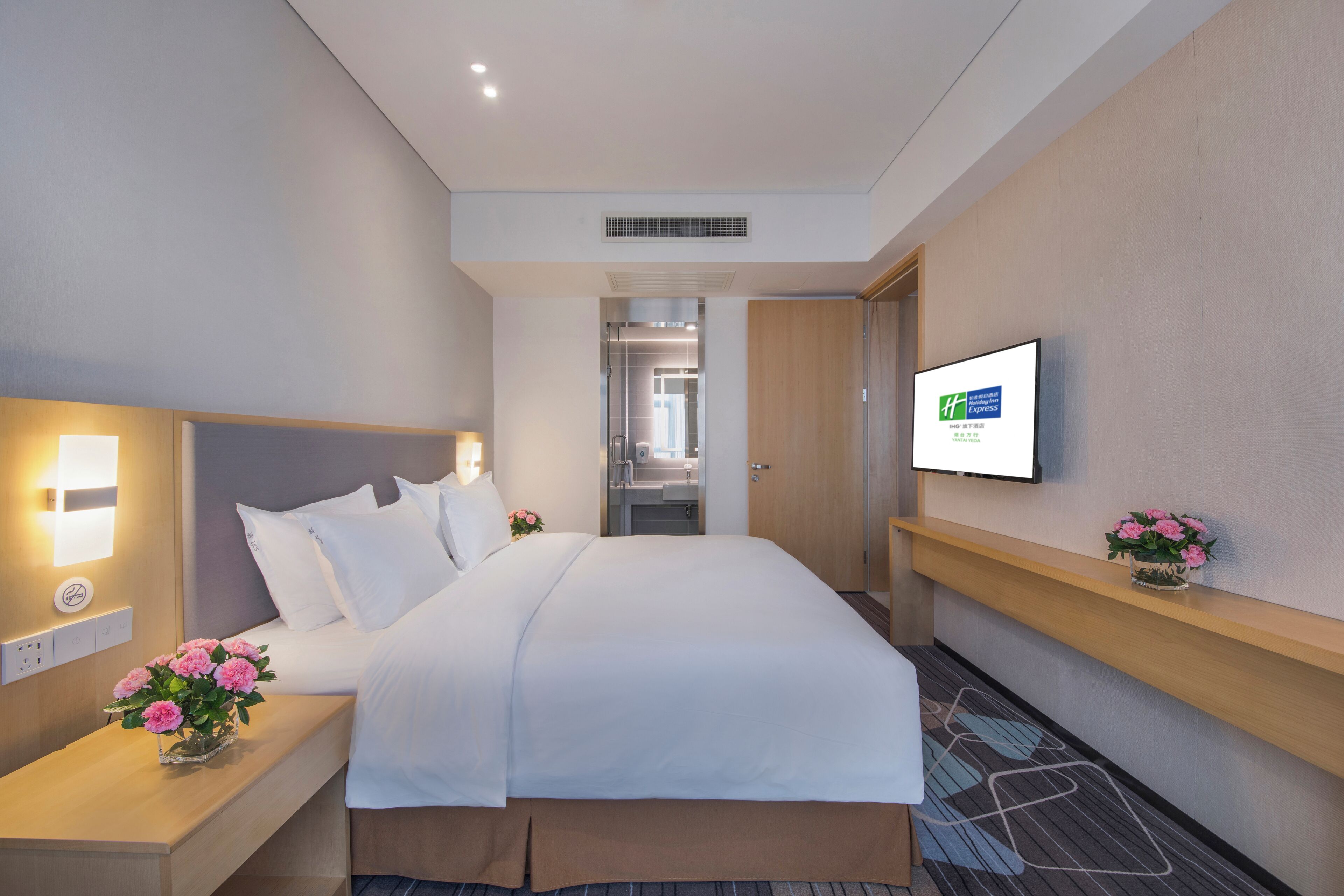 A hotel room with a large bed, bedside tables, a TV, and a small flower arrangement.
