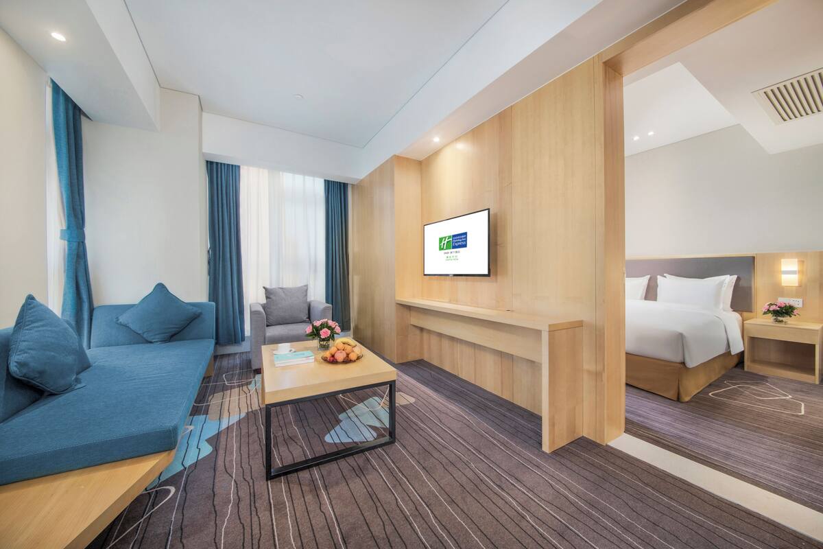 A modern hotel room with a sofa, a coffee table, a bed, a TV, and a wooden accent wall.