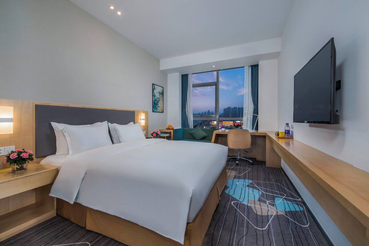 A hotel room with a large bed, a desk, a chair, a TV, and a window with a city view.