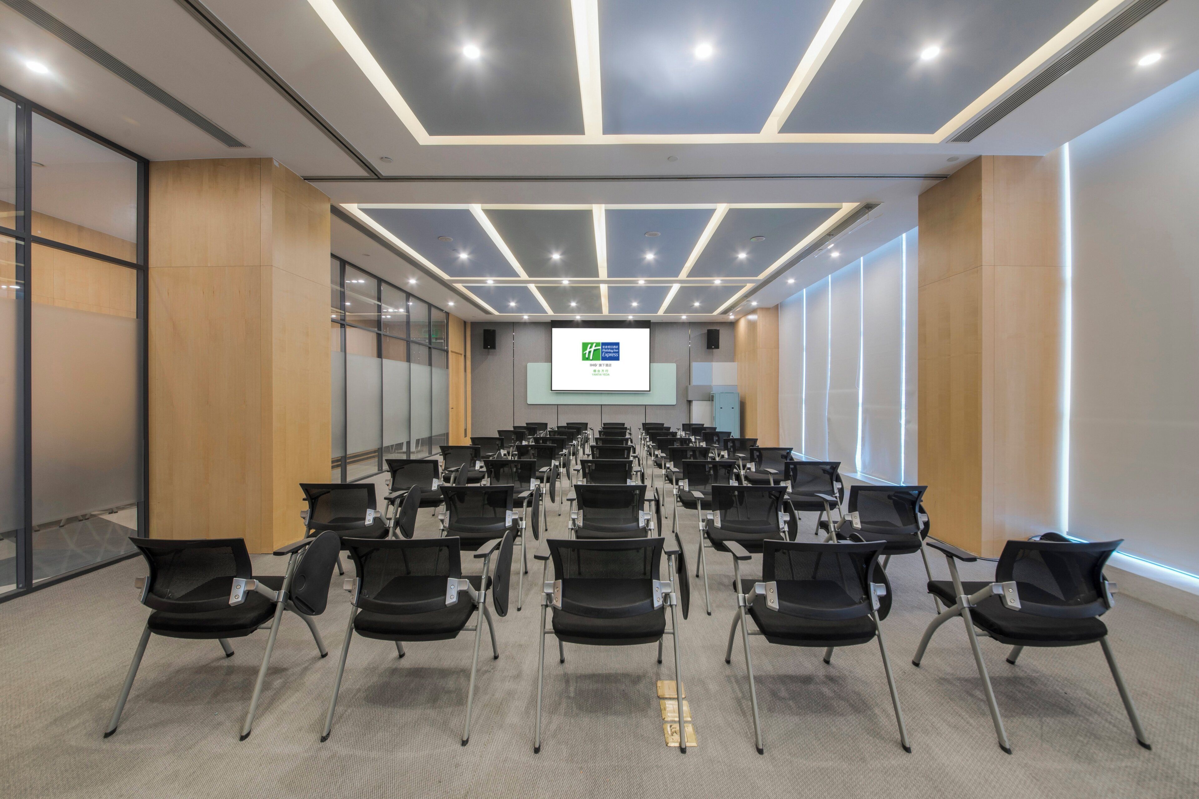 Uma sala de conferências com fileiras de cadeiras pretas, uma tela de projetor na frente e um design moderno no teto.