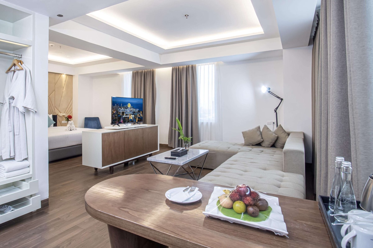 A modern hotel room with a bed, a desk, a sofa, and a dining table with fruit.