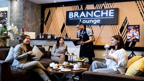 A lounge area with a sign reading "BRANCHCHE Lounge". There are three people seated, one person standing, and a table with various food items and a teapot. - Golden Tulip Essential Makassar (Makassar)