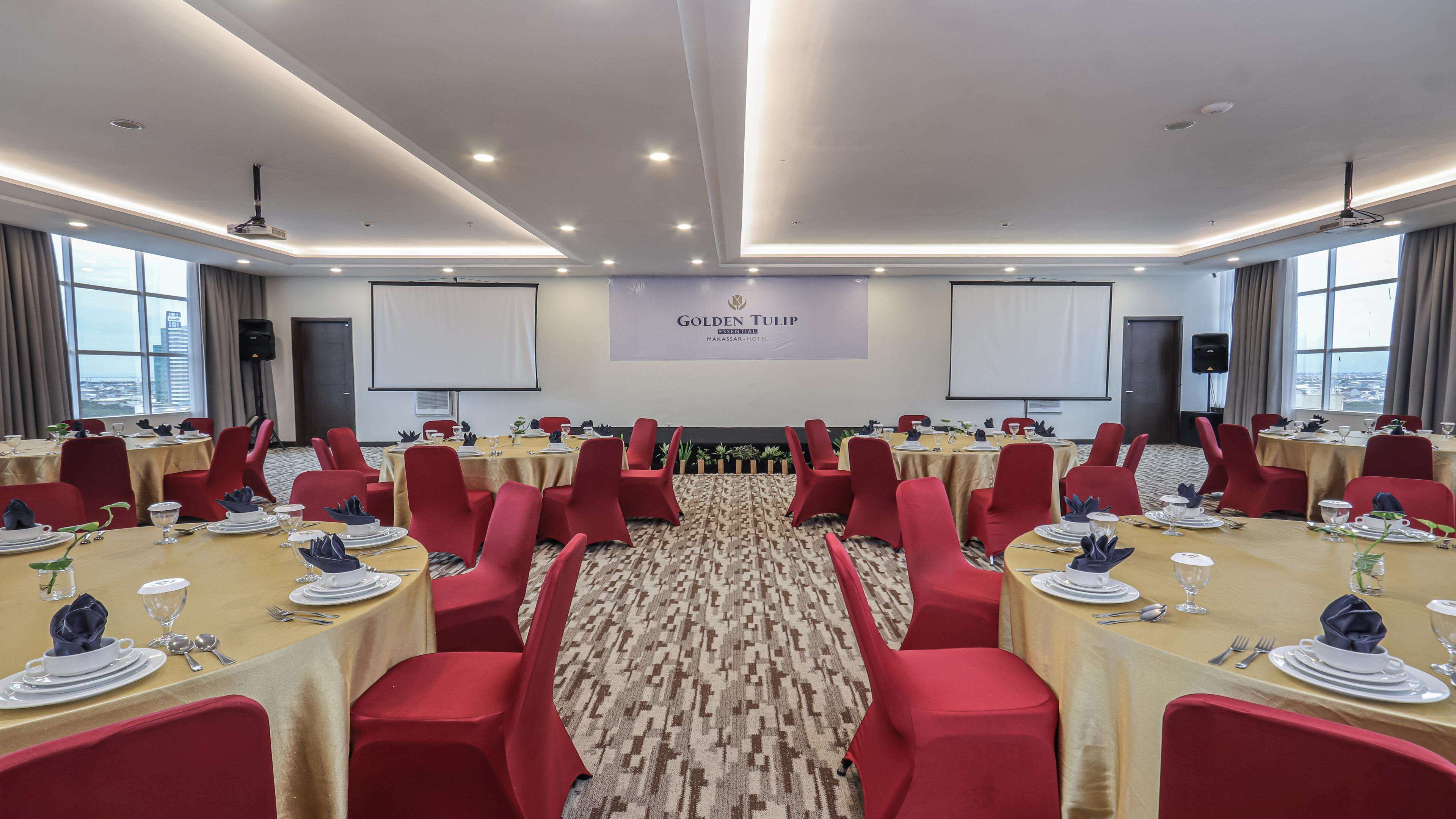 A conference room with round tables, red chairs, and a carpeted floor. There are two large screens on the walls, and the room has a modern ceiling design with recessed lighting.