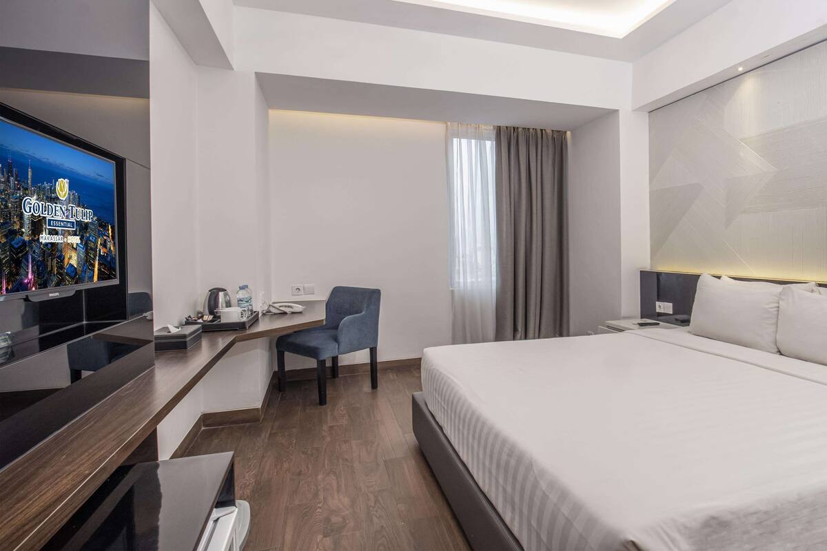 Una habitación de hotel moderna con una cama grande, un escritorio con computadora, una silla y un televisor que muestra un paisaje urbano.