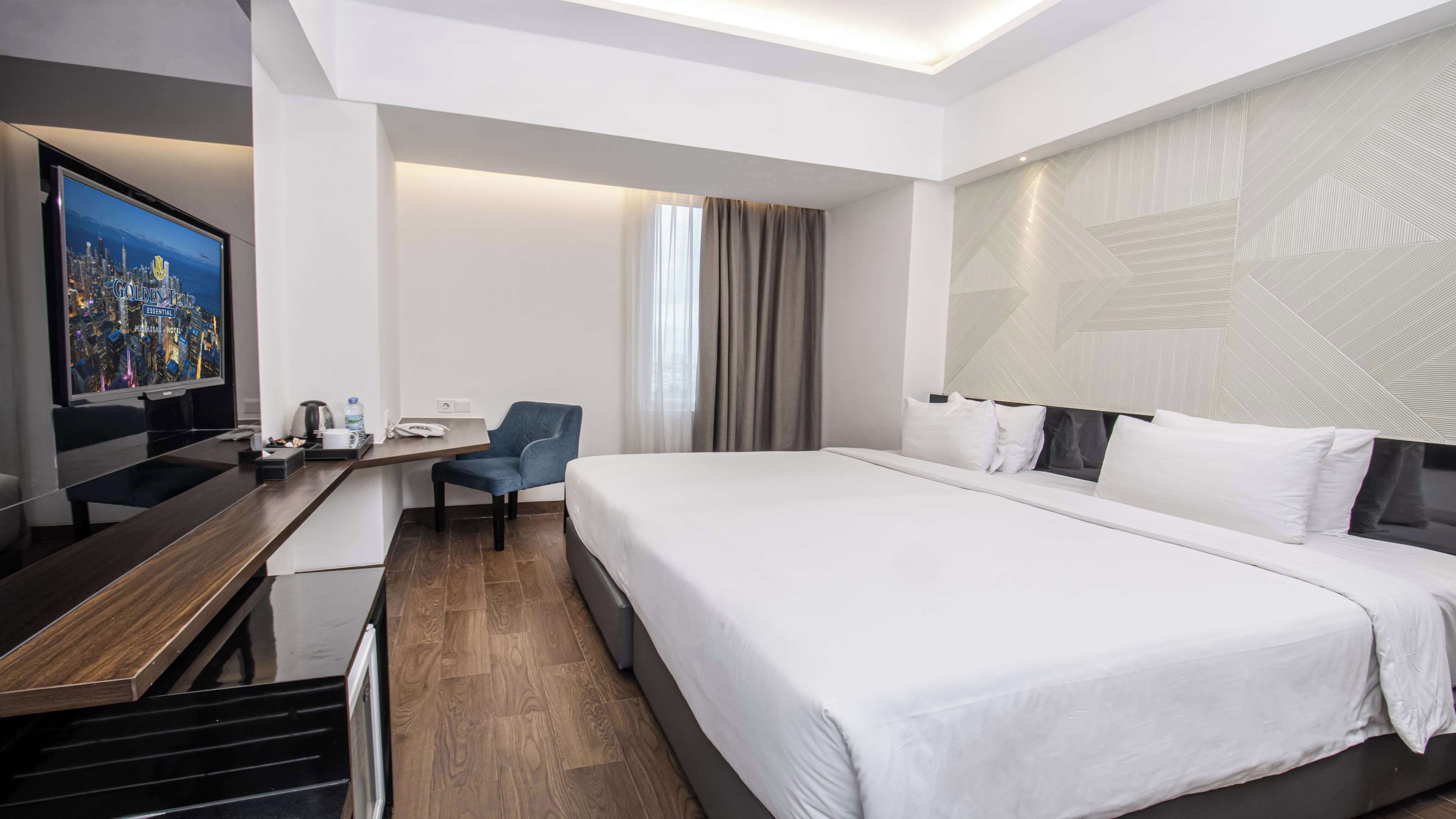 Habitación de hotel moderna con una cama grande, un escritorio con computadora, una silla y un televisor que muestra un paisaje urbano.