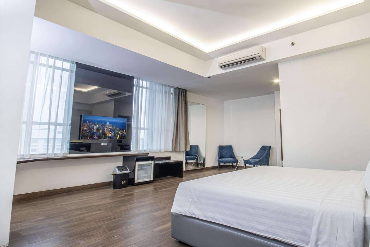 Habitación de hotel moderna con una cama grande, un escritorio con computadora, dos sillas, un espejo amplio y un televisor que muestra un paisaje urbano.
