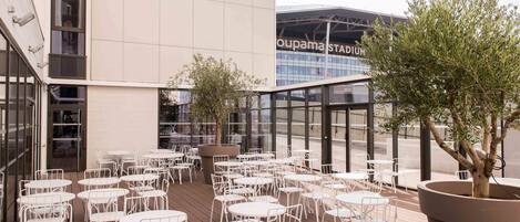 Un espacio al aire libre con mesas y sillas de metal blanco, rodeado por un edificio con amplias ventanas y un letrero que dice "oupmastadium".