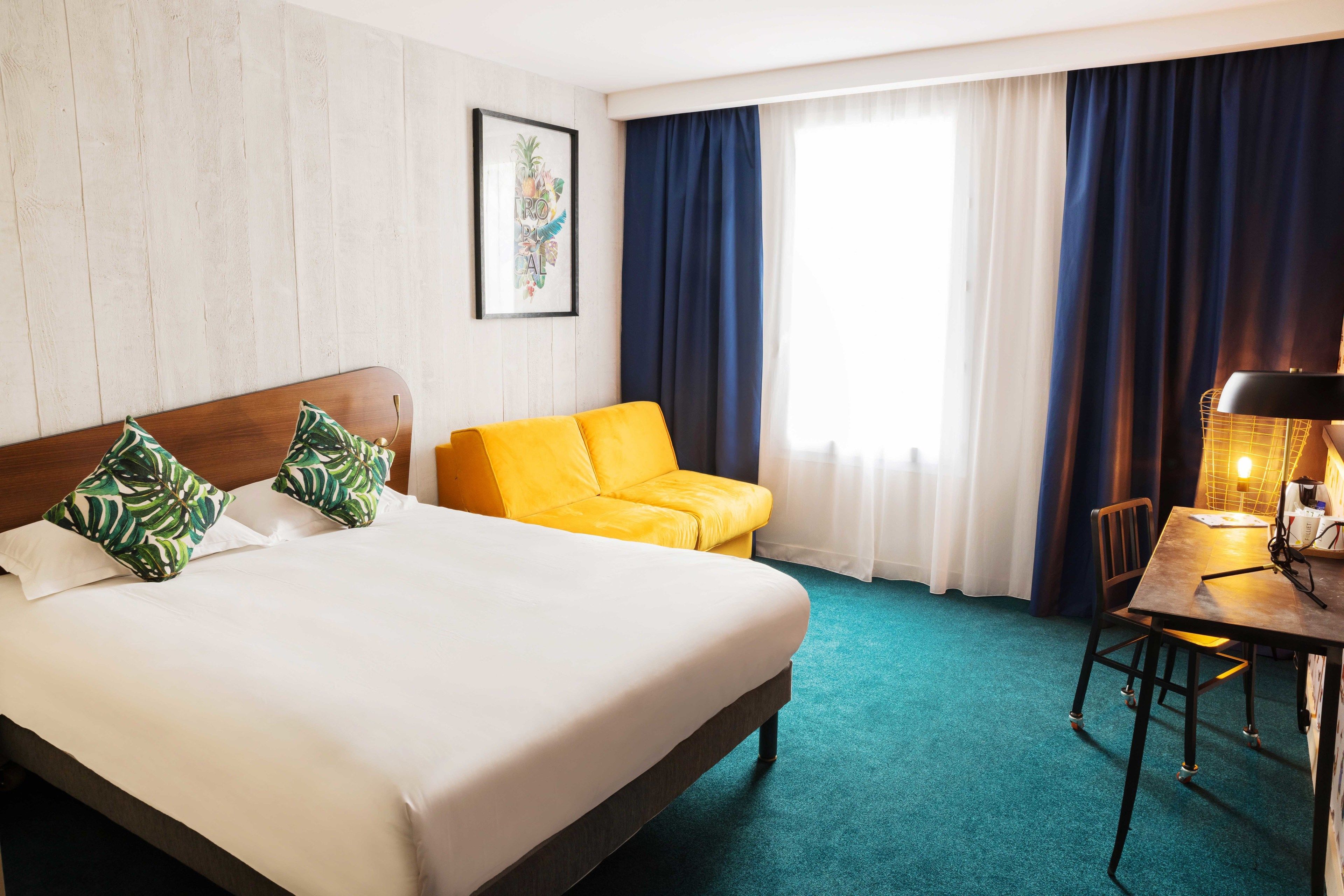 Una habitación de hotel con una cama, un sofá amarillo, un escritorio con lámpara y una ventana con cortinas.