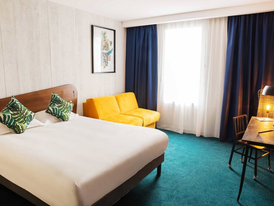 Una habitación de hotel con una cama, un sofá amarillo, un escritorio con lámpara y una ventana con cortinas.