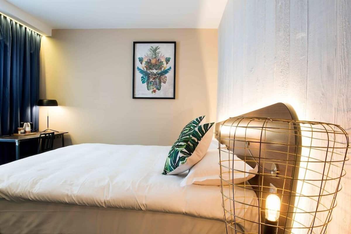 Et hotelværelse med en seng, et skrivebord med lampe, et billede af tropisk kunst på væggen og en usædvanlig lampe med wireframe-design.