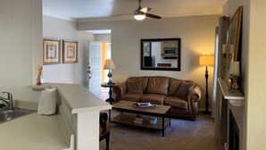 A living room with a brown leather sofa, a coffee table, a ceiling fan, a kitchen area with a microwave, and a fireplace.