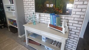 A room with a white brick wall, a large fish tank, a white console table with books, and a small shelf with decorative items. - Turtle Cove Lakefront APT.  10 min from MIS, 40 min to Ann Arbor (Clarklake)