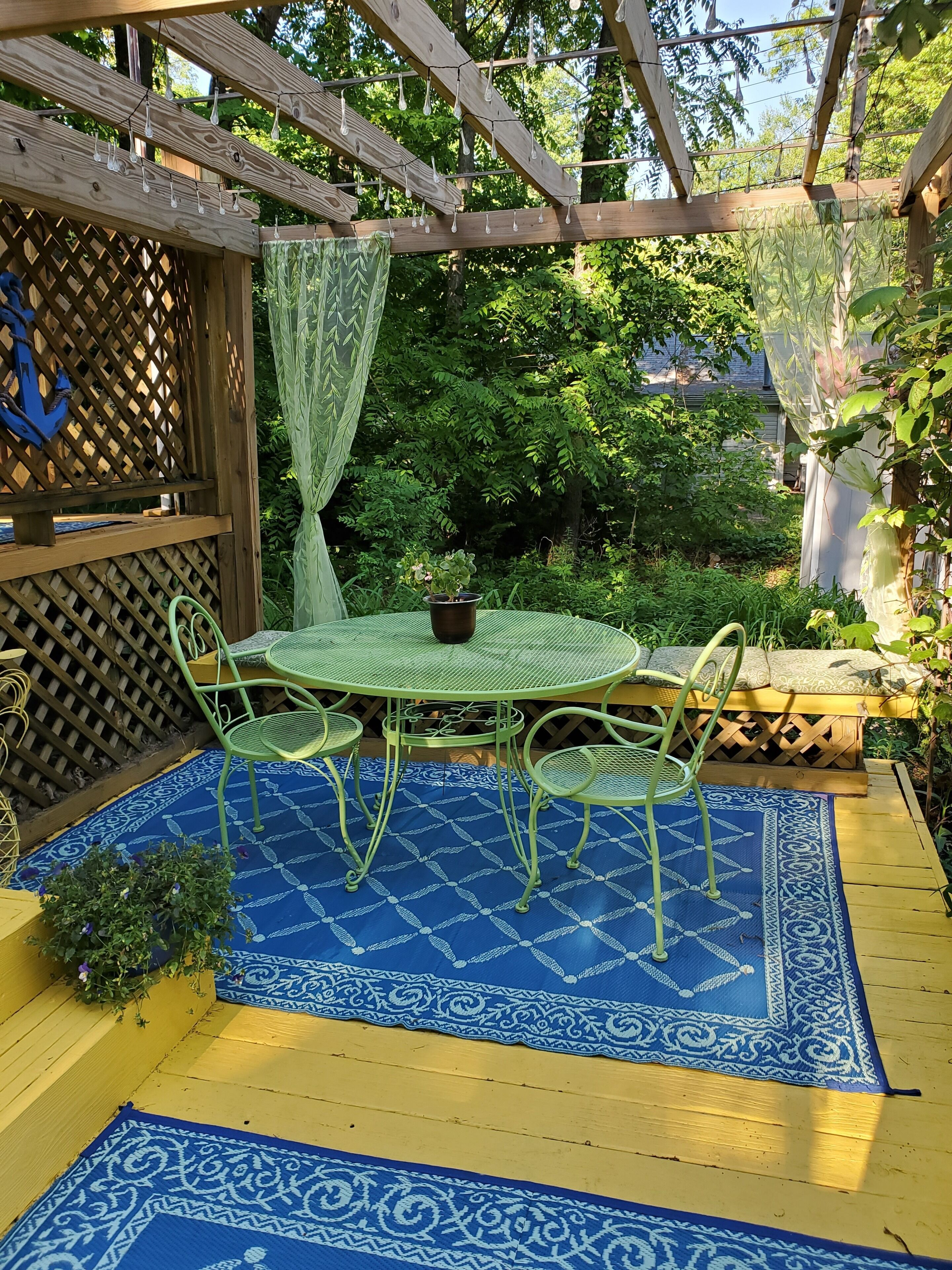 A wooden pergola with string lights, a round table with chairs, and a blue patterned rug.