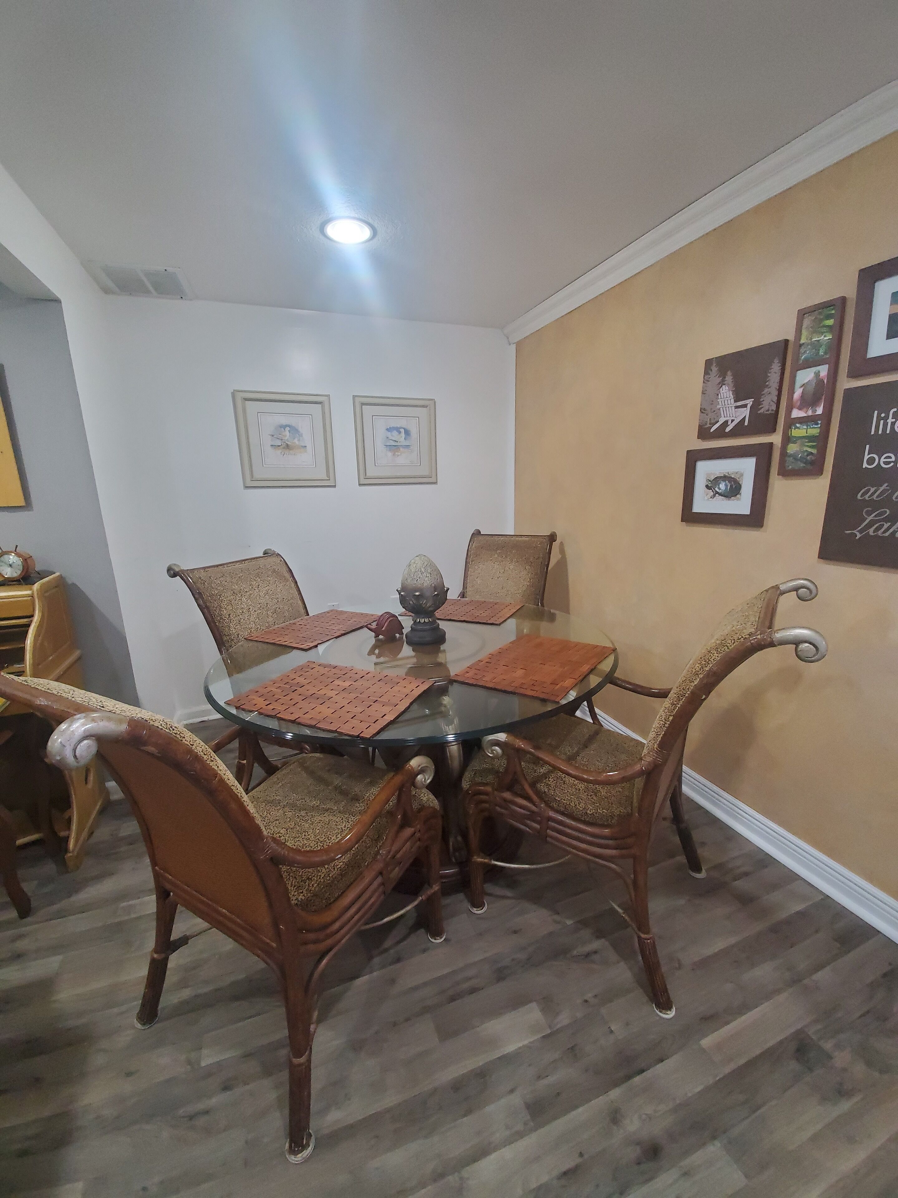 Un coin repas avec une table à plateau de verre entourée de chaises en osier, des tableaux encadrés au mur et une horloge sur le côté gauche.