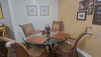 Un coin repas avec une table à plateau de verre entourée de chaises en osier, des tableaux encadrés au mur et une horloge sur le côté gauche.