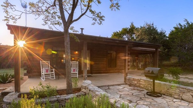 A rustic cabin with a stone patio, a wooden porch, and a rocking chair. - Romantic Log Cabin on Smith Creek with EV Charger (Wimberley)