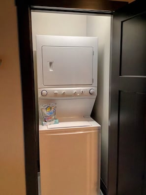 A compact, modern laundry room with a built-in washer and dryer, a small shelf with detergent, and a door with a handle.