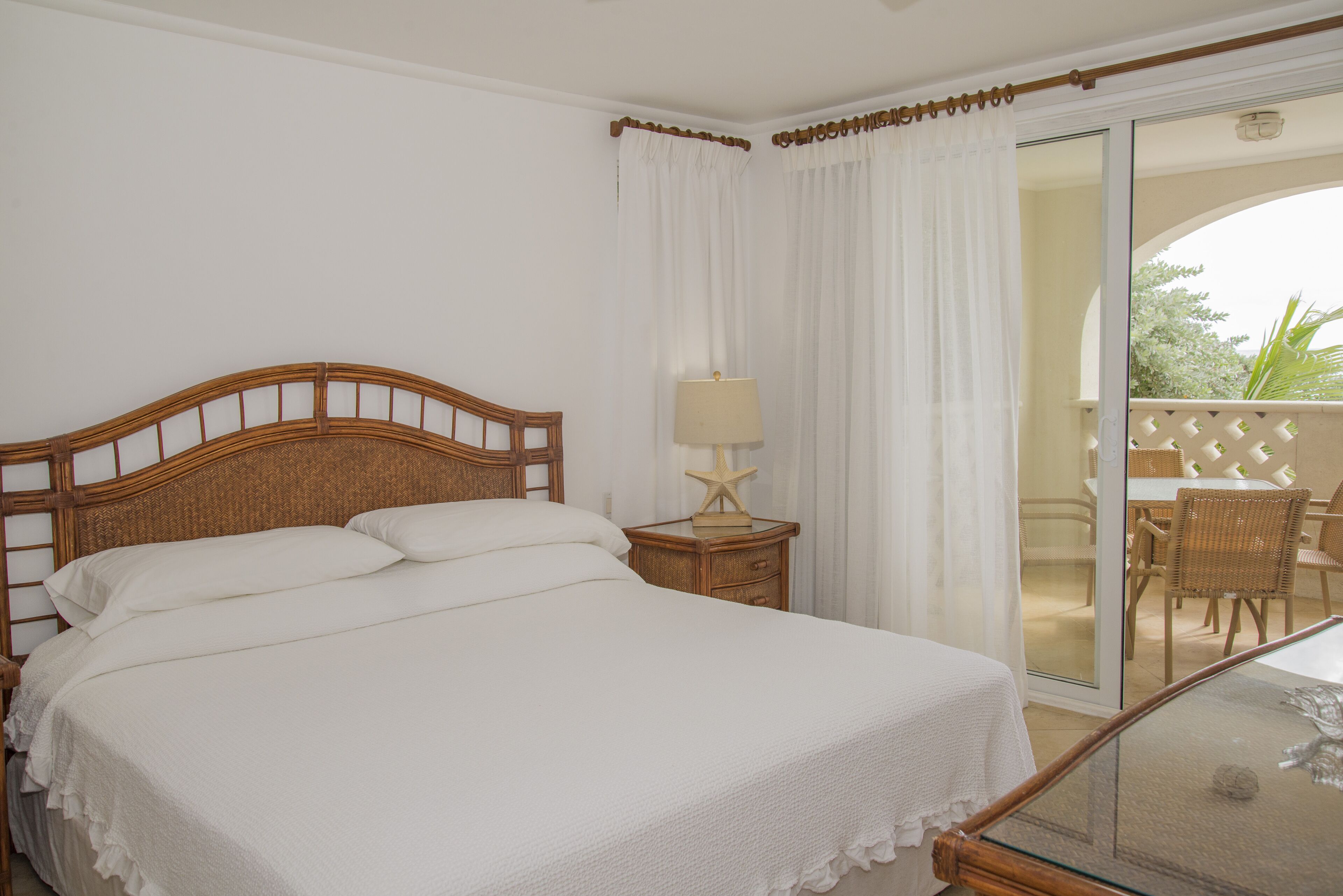 Un dormitorio con cabecera de madera, cama con ropa de cama blanca, mesita de noche con lámpara en forma de estrella y puerta de vidrio que da a un balcón con muebles de mimbre.