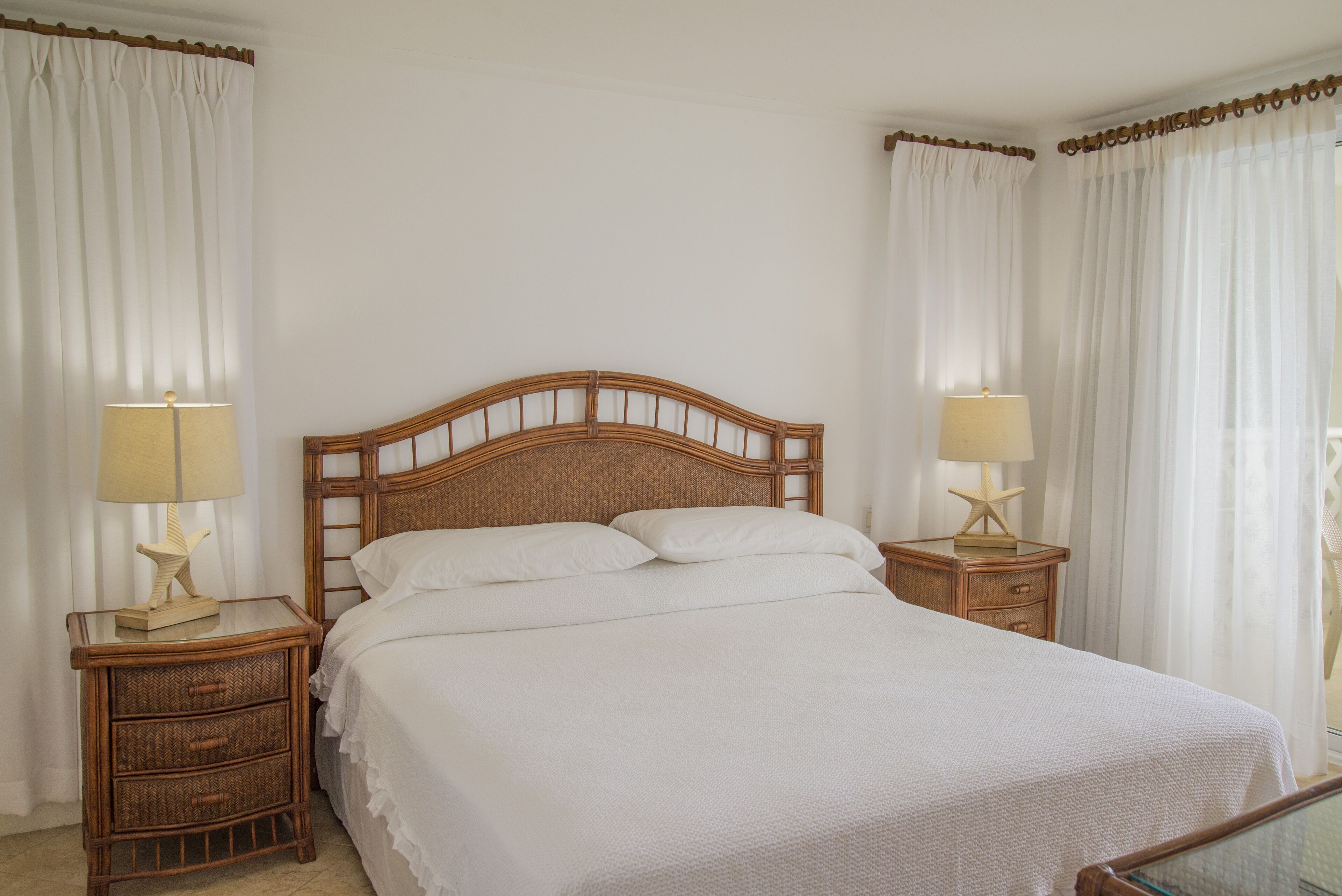 Una cama bien tendida con cabecera de madera, dos mesitas de noche con lámparas y cortinas blancas.