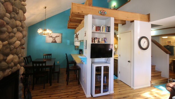 A living room with a stone wall, wooden floor, a dining area with chairs and a table, a bookshelf, a television, and a staircase leading to another level. - Only steps to the slopes!!! (Angel Fire)