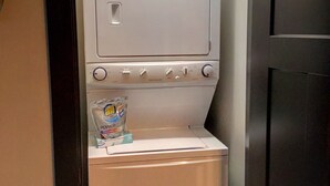 A compact, modern laundry room with a built-in washer and dryer, a small shelf with detergent, and a door with a handle.