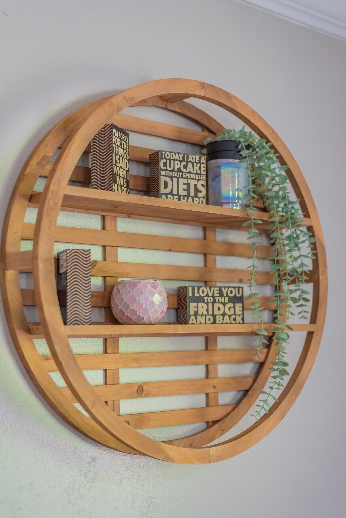 Eine Holz-Wanddekoration mit verschiedenen Gegenständen, darunter eine Cupcake-Box, eine Kerze und ein Schild mit der Aufschrift 'Ich liebe dich bis zum Kühlschrank und wieder zurück'.