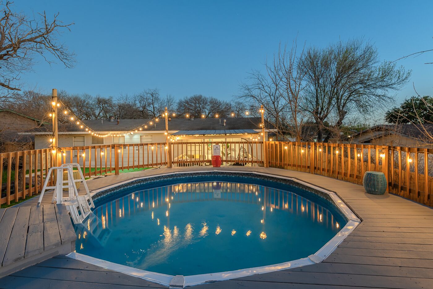 A swimming pool with a wooden deck, surrounded by a wooden fence and string lights.