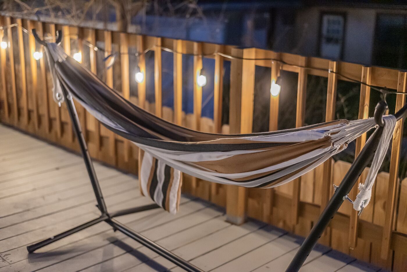 Una hamaca atada a un soporte metálico en una terraza de madera con luces colgantes al fondo.