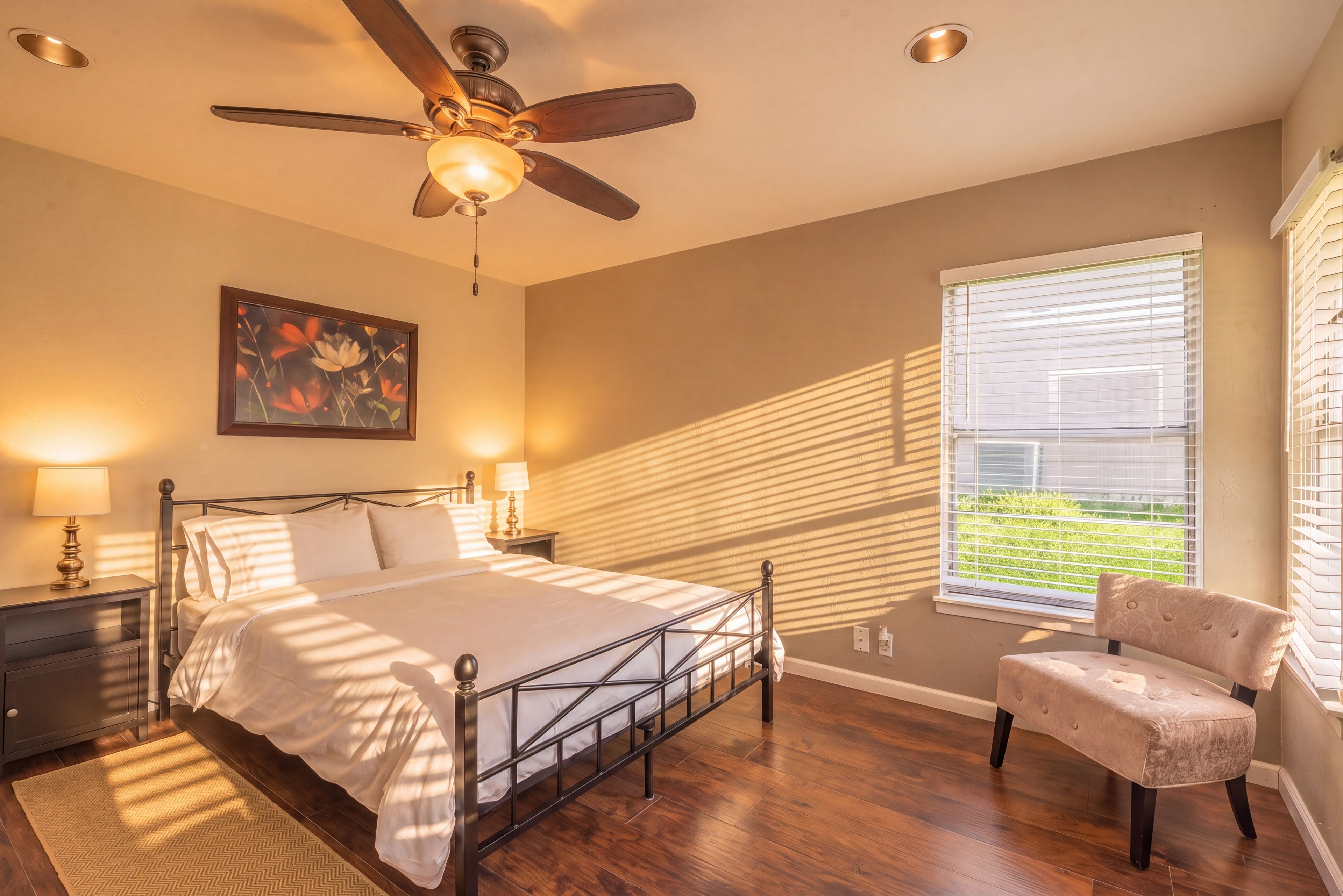A bedroom with a bed, a chair, a ceiling fan, a window with blinds, and a painting on the wall.