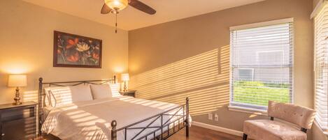 A bedroom with a bed, a chair, a ceiling fan, a window with blinds, and a painting on the wall.