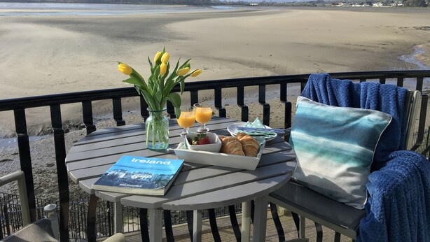 A table with a book titled "Ireland," a vase with yellow flowers, a plate of croissants, and a glass of orange juice on a balcony overlooking a beach and mountains.