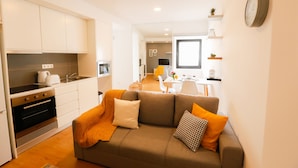 A modern kitchen with white cabinets and a built-in oven, a cozy living area with a brown sofa and orange throw blanket, and a dining area with a white table and chairs. - Historic center of Ponta Delgada - Balcony and Free Wifi (Ponta Delgada)