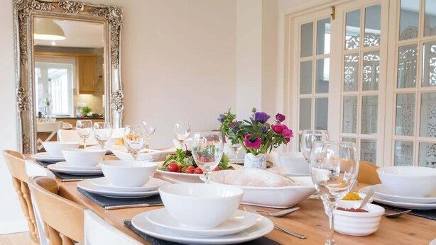 A dining table set for a meal with white plates, bowls, and glasses, a vase with flowers, and a mirror on the wall.
