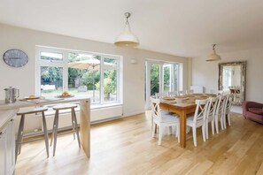 Une cuisine spacieuse avec de grandes fenêtres, une table à manger dressée pour six personnes et une horloge au mur.