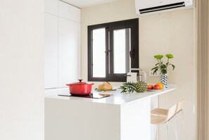 A modern kitchen with white cabinetry, a black-framed window, a red pot, a loaf of bread, a pineapple, and a fruit bowl.
