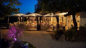 An outdoor seating area with tables and chairs under a covered structure, surrounded by trees and a gravel path. - Bungalow in France with Private Garden (Mauroux)