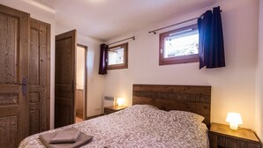 Un dormitorio con cabecera de madera, cama con ropa de cama estampada, mesita de noche con lámpara, ventana con cortinas y una puerta.