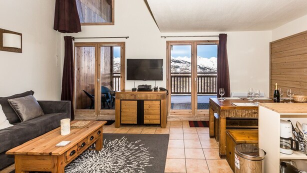 Una sala de estar moderna con un sofá, mesa de centro, televisor y un balcón con vistas a montañas nevadas.