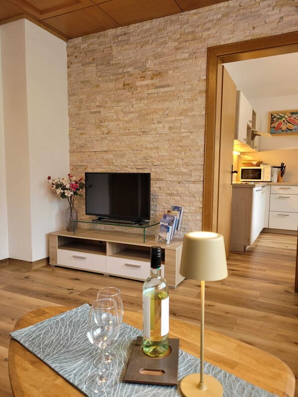 A modern living room with a stone wall, a flat-screen TV, a glass coffee table with wine glasses and a bottle, and a kitchen in the background.