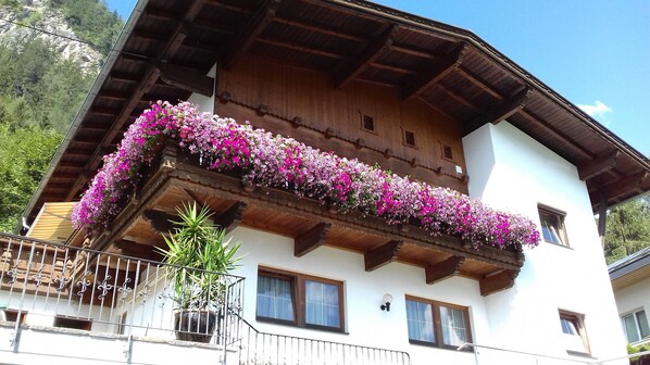 Una cabaña de madera con un balcón decorado con flores rosas, ventanas y un paisaje de montaña al fondo.