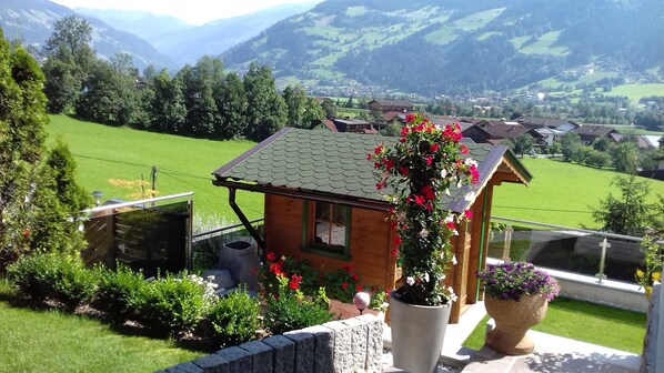 Uma cabana de madeira com telhado inclinado, rodeada por vegetação exuberante e vista para um vale com casas.