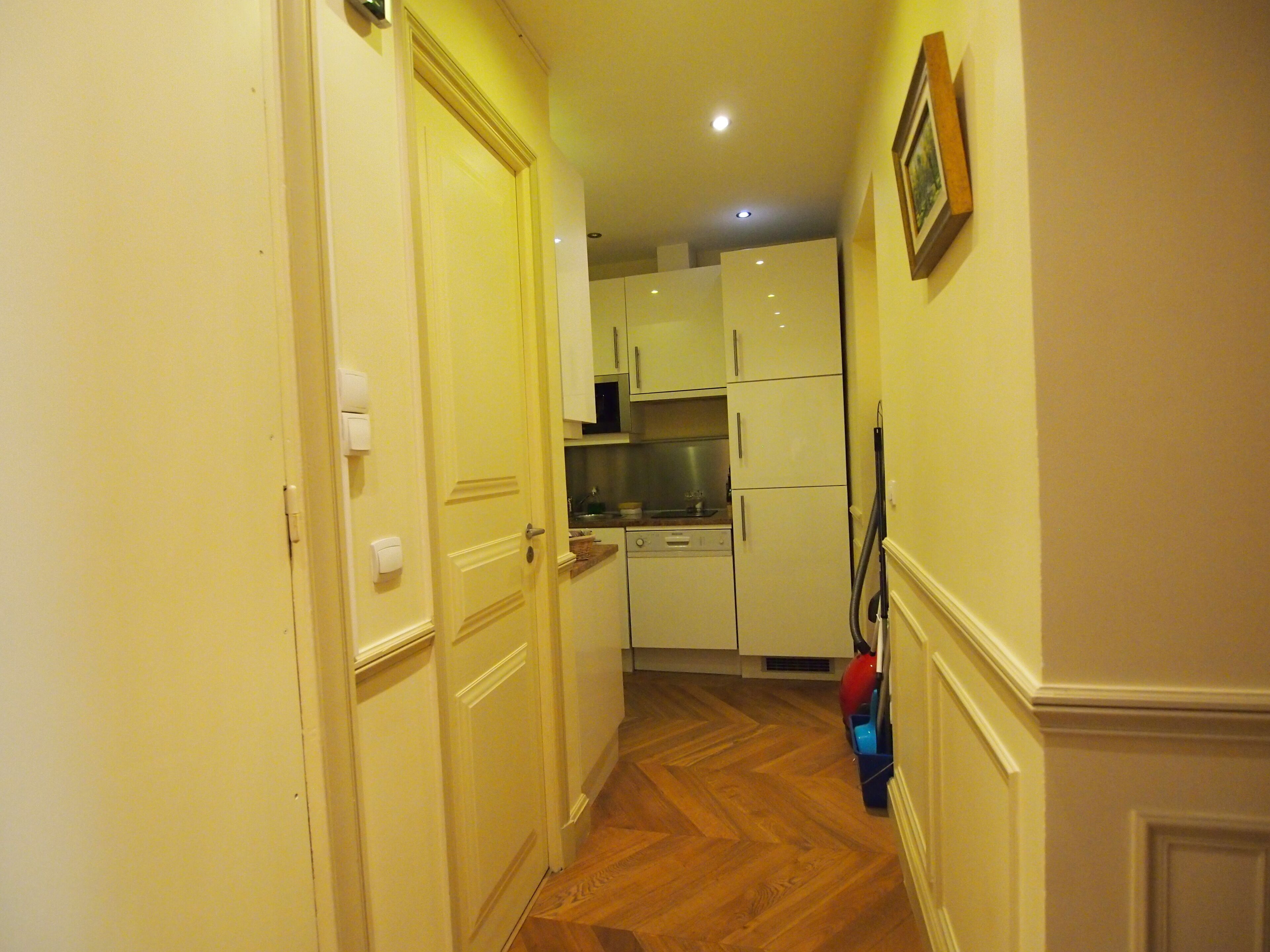A narrow hallway with wooden flooring leading to a kitchen area with white cabinets and a red appliance. - Luxury apartment at the heart of Paris Rent (Paris)