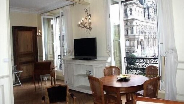A dining area with a chandelier, a television on a cabinet, and a view of a building through a window. - Luxury apartment at the heart of Paris Rent (Paris)