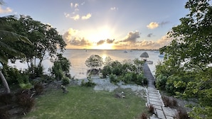 A tropical garden with a wooden deck leading to a beach with boats and a sunset. - Beautiful Beachfront Island Home, Private Dock, Village Transportation Included (Placencia)