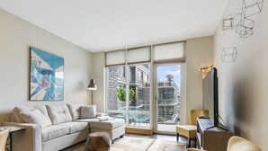 A modern living room with a sofa, a coffee table, a TV, and a large window with a view of buildings. - Pearl Pad: Enjoy River, Shopping, Dining.  Balcony, Garage  and free EV chging! (Portland)