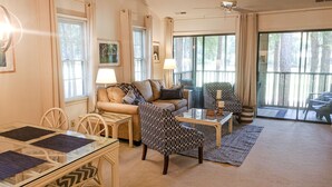 A spacious living room with a sofa, armchairs, a glass coffee table, and a dining table. Large windows provide natural light and a view of the outdoors.